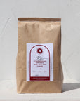 Photo showing packaged up rye flour sitting upright in a brown paper bag with Capital millers sticker on the front. The sticker has all the product information and a burgundy accent colour. White, textured background