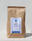 Photo showing packaged up buckwheat flour sitting upright in a brown paper bag with Capital millers sticker on the front. The sticker has all the product information and a turquoise accent colour. White, textured background